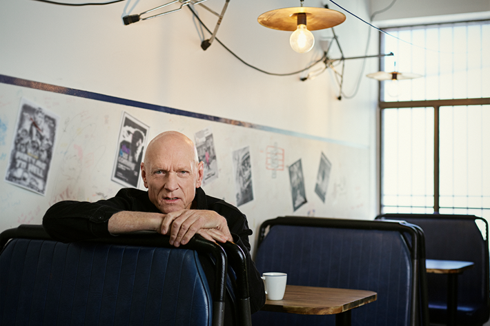 Person leaning on a diner booth with a coffee cup, surrounded by posters and industrial-style lighting.