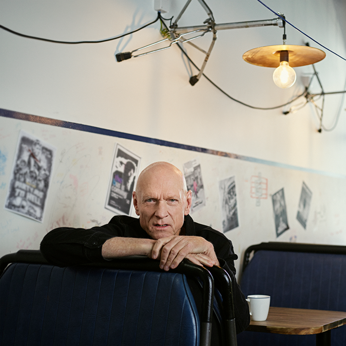 Person leaning on a diner booth with a coffee cup, surrounded by posters and industrial-style lighting.