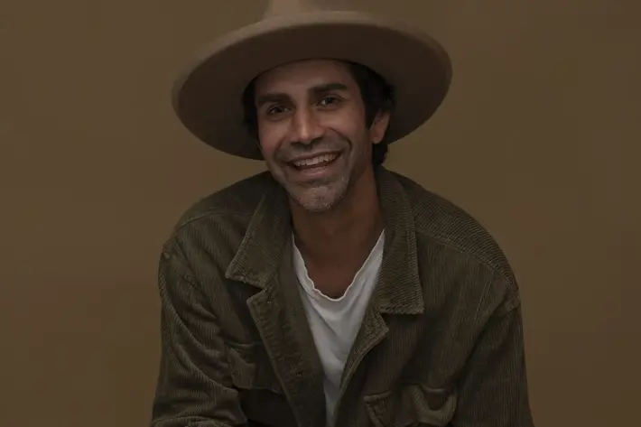 A photo of Brett Lee (Pirritu). A person is seated against a plain brown background, wearing a wide-brimmed hat, a white T-shirt, and a textured brown jacket. Their hands are clasped together and resting on their knees, with dark trousers completing the outfit.