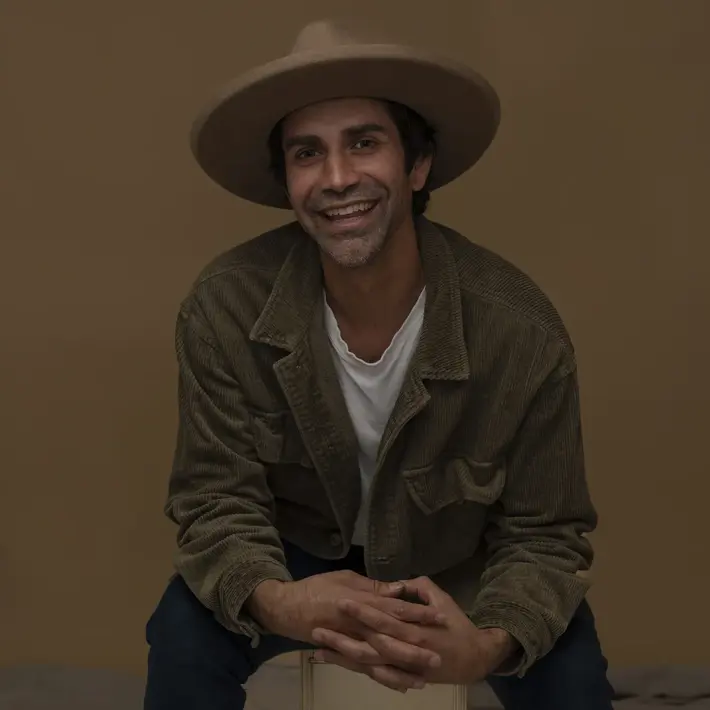 A photo of Brett Lee (Pirritu). A person is seated against a plain brown background, wearing a wide-brimmed hat, a white T-shirt, and a textured brown jacket. Their hands are clasped together and resting on their knees, with dark trousers completing the outfit.