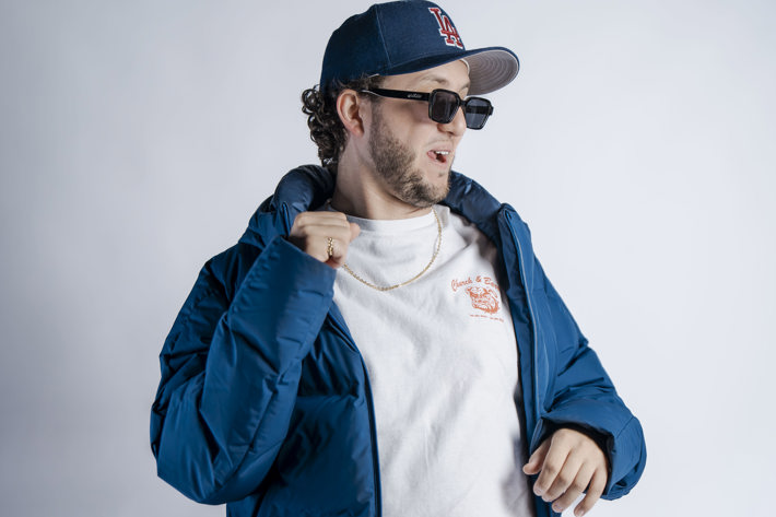 A person in a blue puffer jacket, white t-shirt, and denim jeans, wearing sunglasses and a baseball cap against a light background.
