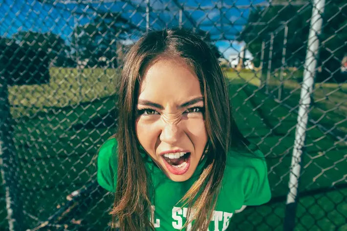 A person with long brown hair stands outdoors in front of a chain-link fence on a sunny day. They are wearing a green T-shirt, and the background shows grass, trees and sporting nets. The lighting creates strong shadows, giving the scene a bright, high‑contrast look.