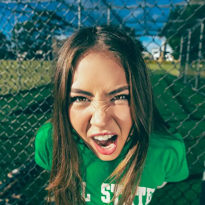 A person with long brown hair stands outdoors in front of a chain-link fence on a sunny day. They are wearing a green T-shirt, and the background shows grass, trees and sporting nets. The lighting creates strong shadows, giving the scene a bright, high‑contrast look.