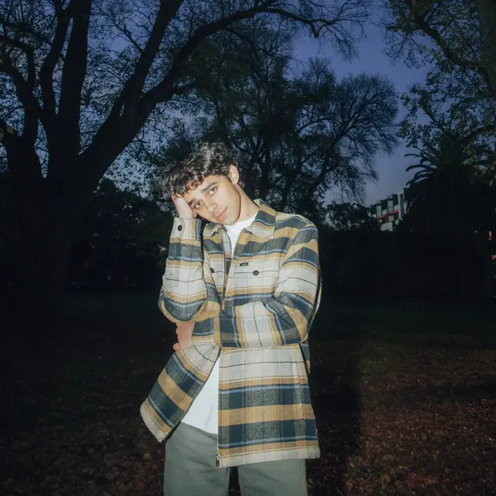 A person stands outdoors in a park during dusk, wearing a long-sleeved, yellow and grey checked flannel jacket over a white shirt. Tall trees with bare branches frame the background against a deep blue evening sky, with a few scattered leaves visible. Some buildings and palm trees can be seen in the distance, partially obscured by shadows. The lighting highlights the jacket’s texture and colours, creating a moody, atmospheric scene.