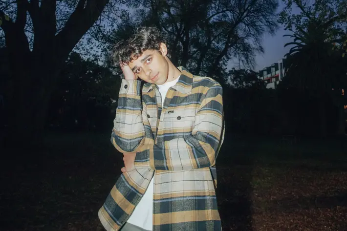 A person stands outdoors in a park during dusk, wearing a long-sleeved, yellow and grey checked flannel jacket over a white shirt. Tall trees with bare branches frame the background against a deep blue evening sky, with a few scattered leaves visible. Some buildings and palm trees can be seen in the distance, partially obscured by shadows. The lighting highlights the jacket’s texture and colours, creating a moody, atmospheric scene.