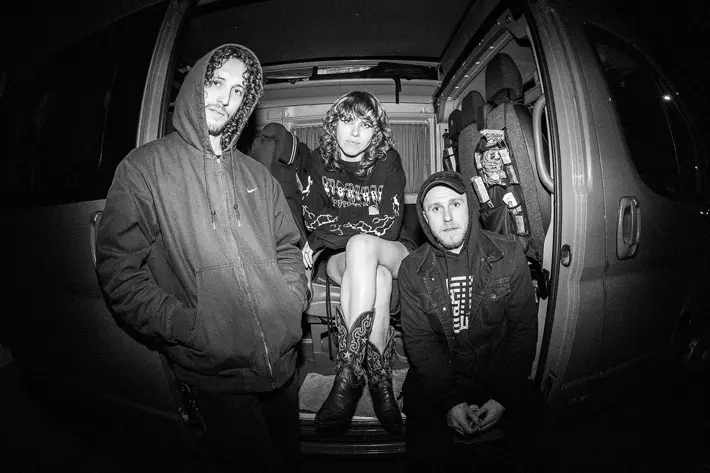 Three people wearing hooded jackets and casual clothing sit and kneel inside an open van at night. One person in the centre is seated with legs crossed, wearing patterned boots and a graphic hoodie. The van interior shows seats and various items stored on the side. The image is in black and white, creating a moody, high-contrast atmosphere. Photo by Emmanuel Poteau
