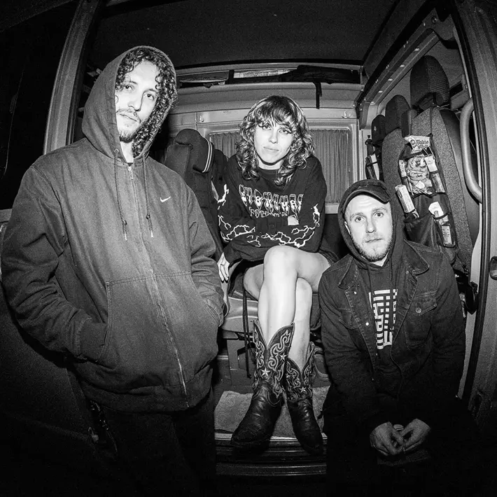 Three people wearing hooded jackets and casual clothing sit and kneel inside an open van at night. One person in the centre is seated with legs crossed, wearing patterned boots and a graphic hoodie. The van interior shows seats and various items stored on the side. The image is in black and white, creating a moody, high-contrast atmosphere. Photo by Emmanuel Poteau