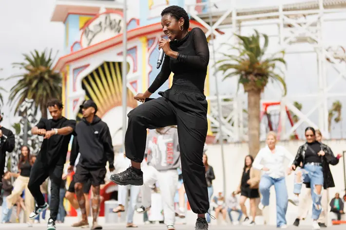 A person dressed in black clothing is performing a dynamic dance move while holding a microphone in an outdoor setting. Behind them, several other individuals are also dancing, creating an energetic group performance. The background features a colourful amusement park entrance with palm trees and a roller coaster structure, suggesting a lively public event or festival atmosphere.