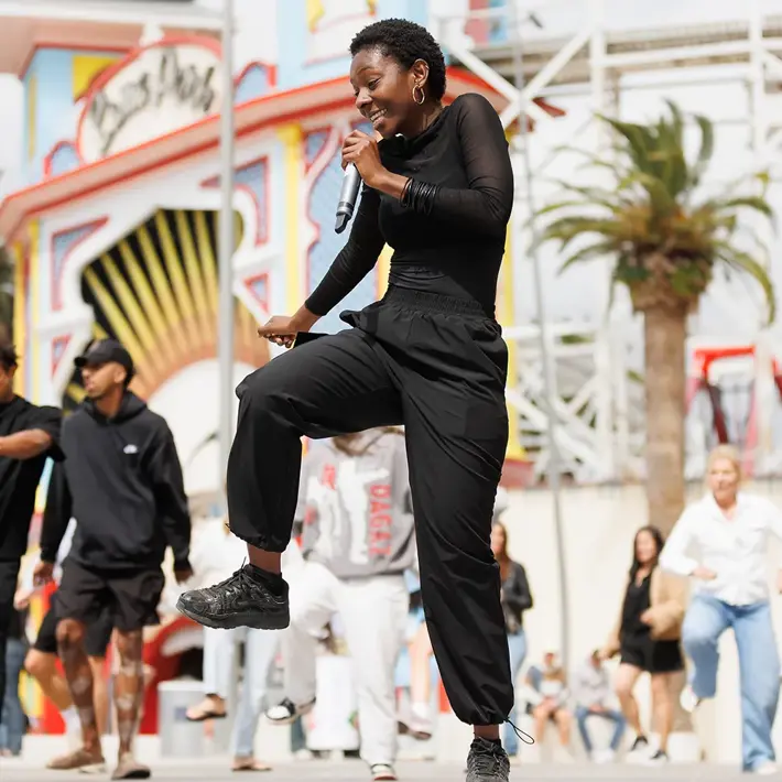 A person dressed in black clothing is performing a dynamic dance move while holding a microphone in an outdoor setting. Behind them, several other individuals are also dancing, creating an energetic group performance. The background features a colourful amusement park entrance with palm trees and a roller coaster structure, suggesting a lively public event or festival atmosphere.