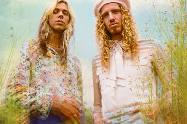 Two individuals with long, curly hair wearing vintage-inspired outfits in a dreamy outdoor setting surrounded by wildflowers.