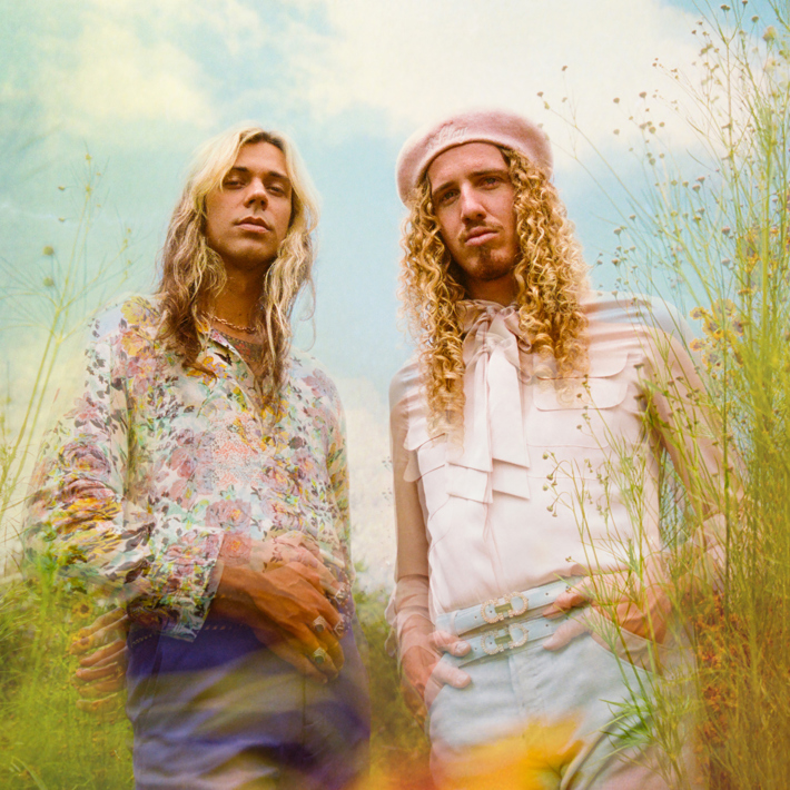 Two individuals with long, curly hair wearing vintage-inspired outfits in a dreamy outdoor setting surrounded by wildflowers.