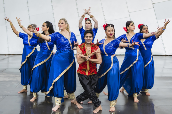 A group of dancers in blue and one in red pose outdoors in front of a large, metallic architectural structure.