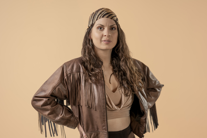 A woman with long hair wearing a headscarf and a fringed leather jacket stands against a beige background.