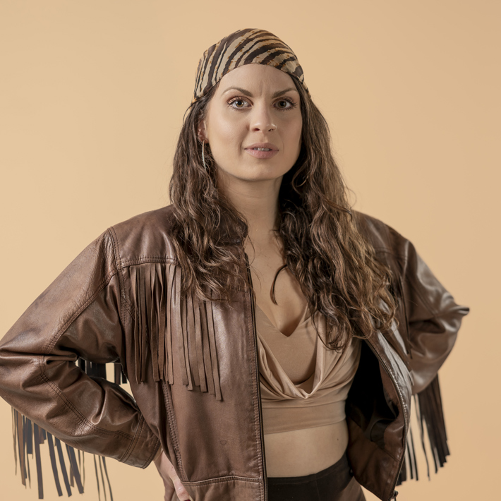 A woman with long hair wearing a headscarf and a fringed leather jacket stands against a beige background.