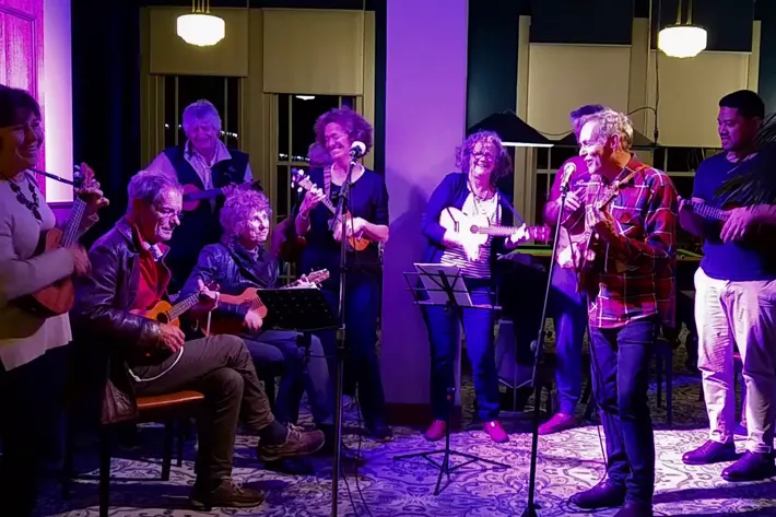 A group of musicians perform indoors under purple stage lighting. They are playing ukuleles and similar string instruments, with some standing and others seated on chairs. Music stands with sheet music are positioned in front of the performers, and microphones are set up for amplification. The background includes large windows, pendant lights, and patterned carpet, creating an intimate live music setting.