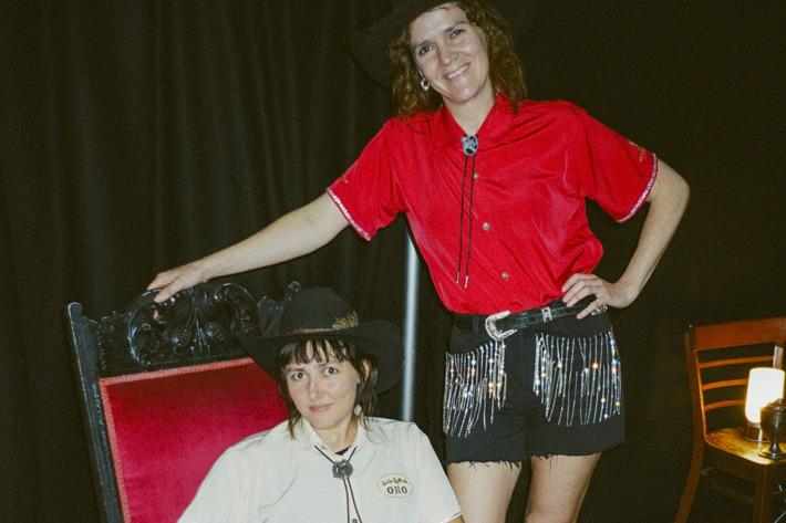 Two individuals dressed in cowboy-inspired outfits pose confidently. One sits on a red velvet chair, while the other stands beside it, both wearing hats and boots.