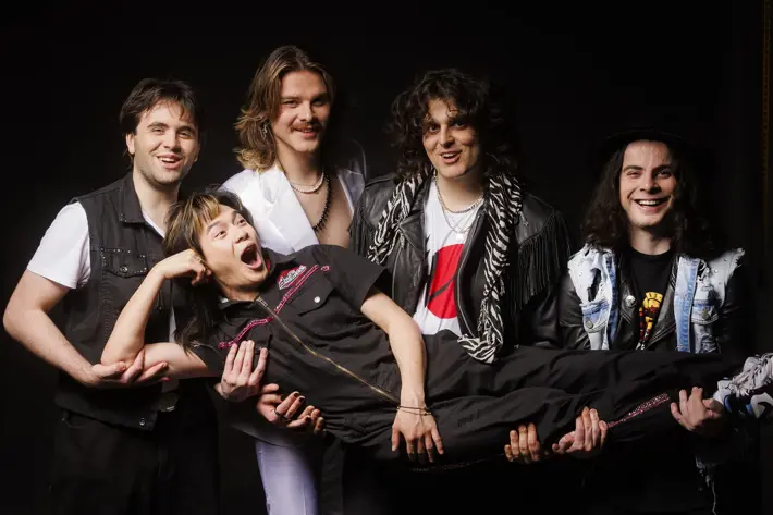 A group photo of five band members posing playfully against a black background. Four members are standing and smiling while holding up the fifth member horizontally across their arms. Everyone looks happy and energetic, dressed in rock-inspired outfits with bright expressions.