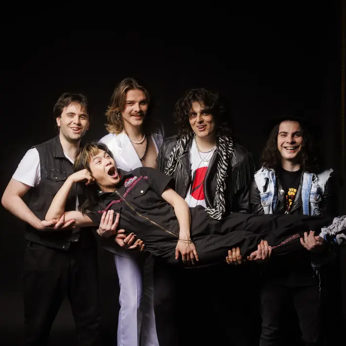 A group photo of five band members posing playfully against a black background. Four members are standing and smiling while holding up the fifth member horizontally across their arms. Everyone looks happy and energetic, dressed in rock-inspired outfits with bright expressions.