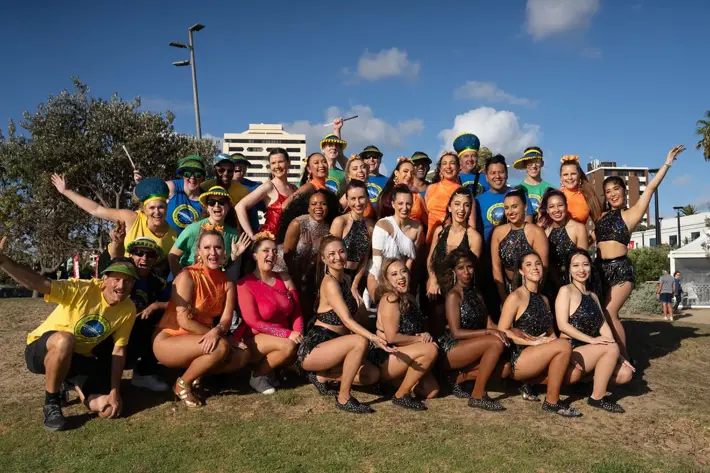 A large group of performers pose outdoors on a grassy area with buildings and palm trees in the background. They wear vibrant costumes, including sequined black dance outfits and colourful shirts in yellow, green, orange, and pink, some featuring Brazilian-themed designs. Several participants accessorise with feathered headpieces and wristbands, and the group is arranged in a dynamic, celebratory pose under a bright blue sky.