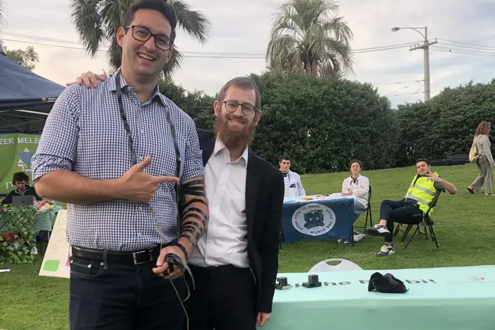 Two individuals stand outdoors near a table with a green cloth that reads “Ask the Rabbi!” in bold text. One person is wearing a checkered shirt and has tefillin wrapped around an arm, while the other is dressed in a dark suit. The setting includes grass, palm trees, and people seated at tables in the background, suggesting a community or cultural event.
