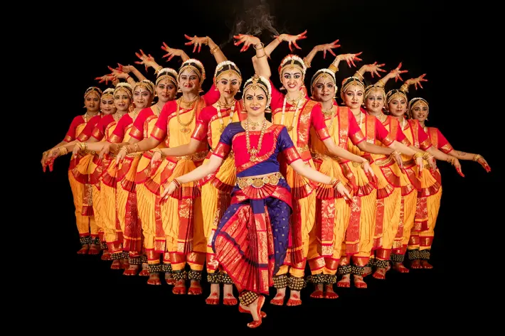 A group of performers in traditional Indian attire pose in a symmetrical formation against a black background. They wear vibrant costumes featuring yellow and red garments with ornate gold detailing, while the central performer is dressed in blue and red. Their arms are raised in expressive, coordinated positions, showcasing a classical dance style.