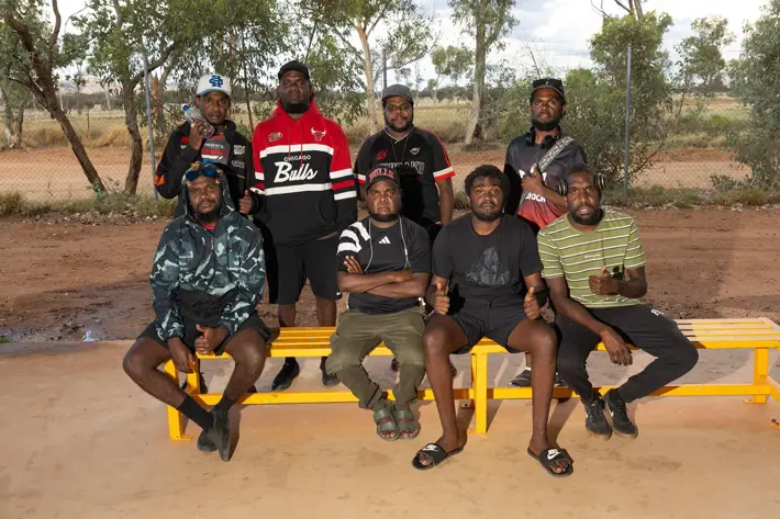 A group of seven people sit and stand around bright yellow benches outdoors on a dirt surface. They are dressed in casual sportswear, including striped shirts, hoodies, and branded jackets. Behind them is a wire fence and scattered trees under a partly cloudy sky.
