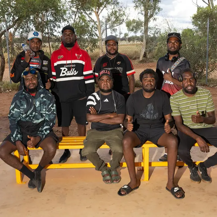 A group of seven people sit and stand around bright yellow benches outdoors on a dirt surface. They are dressed in casual sportswear, including striped shirts, hoodies, and branded jackets. Behind them is a wire fence and scattered trees under a partly cloudy sky.