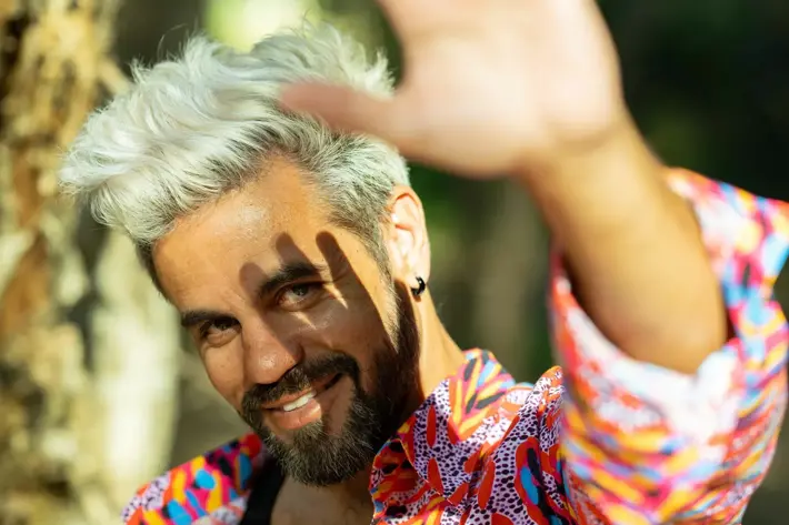 Boox Kid, wearing a bright red, yellow and blue shirt, looking at the camera and smiling. His hand is outstretched in front, casting a shadow of his hand across his face.
