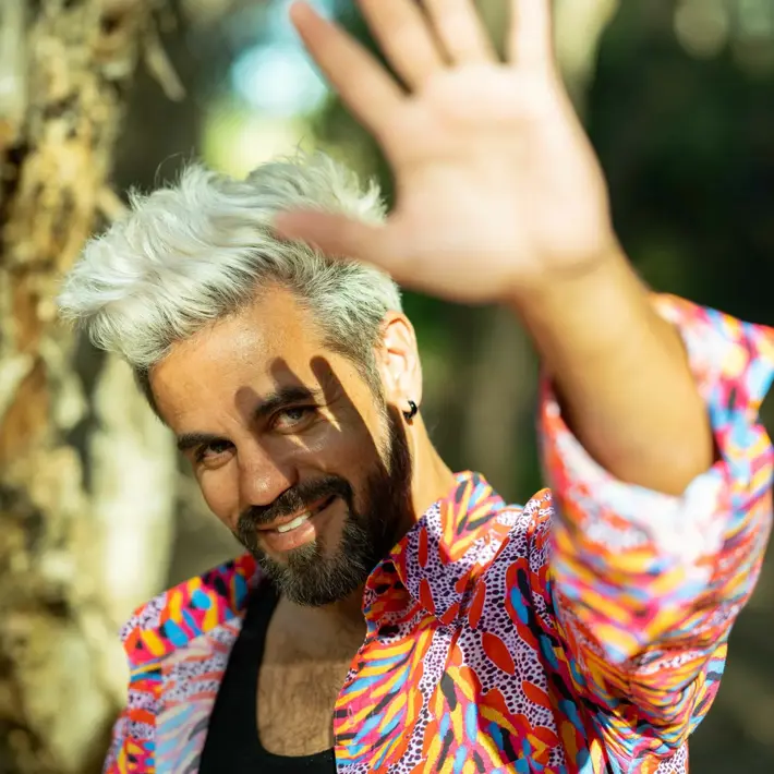 Boox Kid, wearing a bright red, yellow and blue shirt, looking at the camera and smiling. His hand is outstretched in front, casting a shadow of his hand across his face.