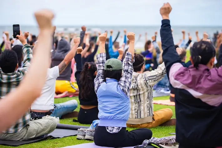 A large group of people sitting on mats outdoors with arms raised in unison, facing towards the ocean. The scene includes individuals wearing casual clothing such as jackets, caps, and patterned shirts. The grassy area is filled with participants, and the background shows a cloudy sky and water, suggesting a coastal setting.