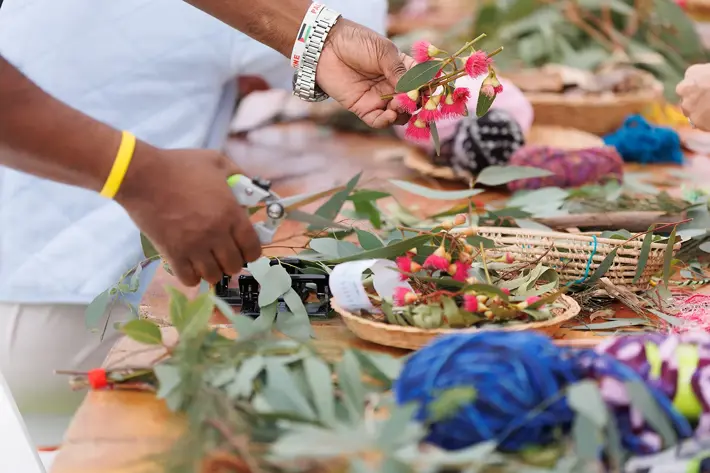 Hands are arranging native Australian flora, including eucalyptus leaves and pink blossoms, on a wooden table. Scissors and woven baskets filled with greenery and flowers are visible, along with colourful yarn in shades of blue and purple. The scene suggests a creative activity such as making bush bouquets or floral arrangements using natural materials.
