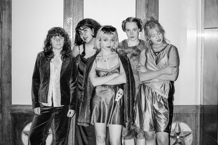 The image features a group of people posing for a photo indoors. They are dressed in various styles, showcasing different clothing and footwear. The photo is in black and white, highlighting their faces as they stand together against a wall.