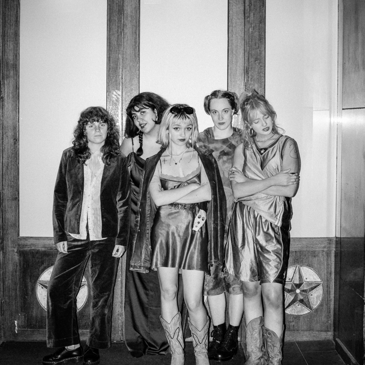 The image features a group of people posing for a photo indoors. They are dressed in various styles, showcasing different clothing and footwear. The photo is in black and white, highlighting their faces as they stand together against a wall.