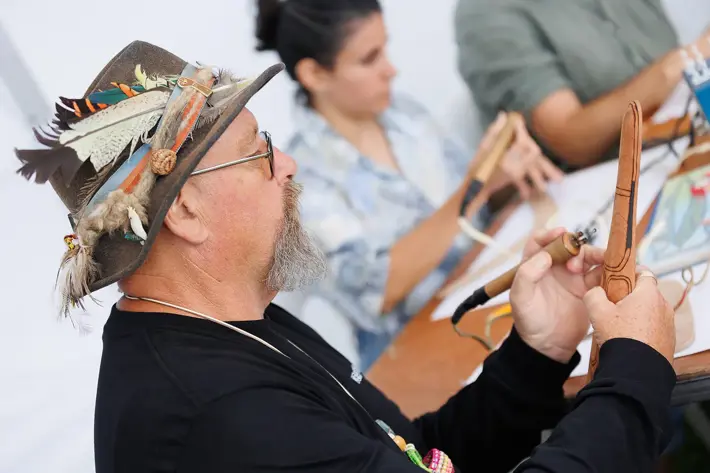 A person is seated at a table, carefully working on a wooden craft piece using a pyrography tool. The individual is wearing a hat decorated with feathers and natural materials. In the background, others are also engaged in similar woodcraft activities, with tools and materials spread across the table. The setting suggests a hands-on workshop focused on traditional or artistic woodcraft techniques.