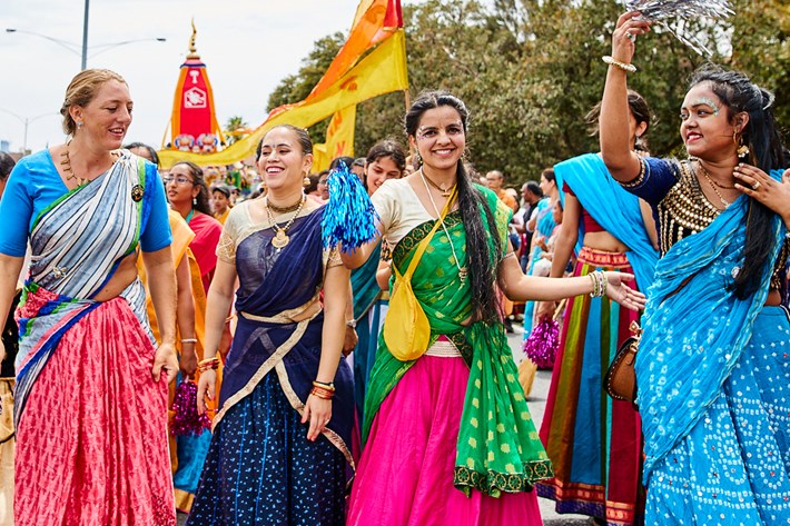 Four women standing side-by-side dancing in front of a large crowd during an outdoor festival while wearing muti-coloured traditional Indian garmets.