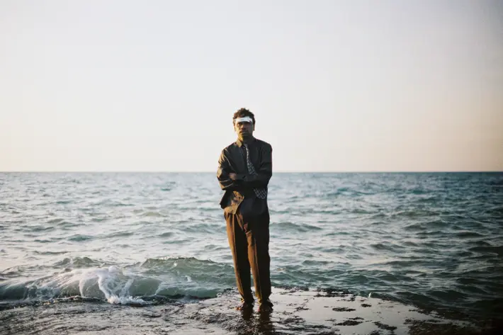 A person is standing on a rocky edge by the sea, with gentle waves washing against the shore. The individual is dressed in a suit with a patterned tie, arms crossed, and facing forward. The background shows a calm ocean under a clear sky with soft, natural lighting, creating a serene and minimalist coastal scene.