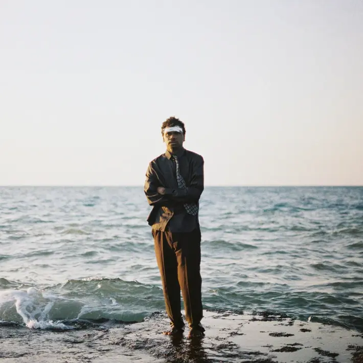 A person is standing on a rocky edge by the sea, with gentle waves washing against the shore. The individual is dressed in a suit with a patterned tie, arms crossed, and facing forward. The background shows a calm ocean under a clear sky with soft, natural lighting, creating a serene and minimalist coastal scene.