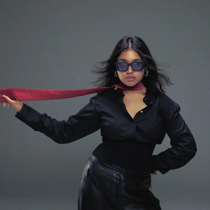 A person poses against a plain grey backdrop wearing a black button-up shirt tucked into black leather trousers. A red striped tie is wrapped loosely around the neck and extends outward, held in one hand, creating a sense of movement. The outfit contrasts sharply with the neutral background, giving the image a bold, stylish, and dynamic feel.