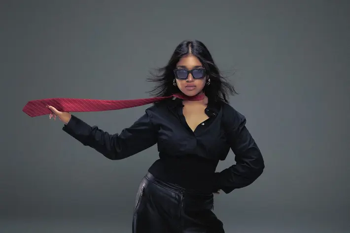 A person poses against a plain grey backdrop wearing a black button-up shirt tucked into black leather trousers. A red striped tie is wrapped loosely around the neck and extends outward, held in one hand, creating a sense of movement. The outfit contrasts sharply with the neutral background, giving the image a bold, stylish, and dynamic feel.
