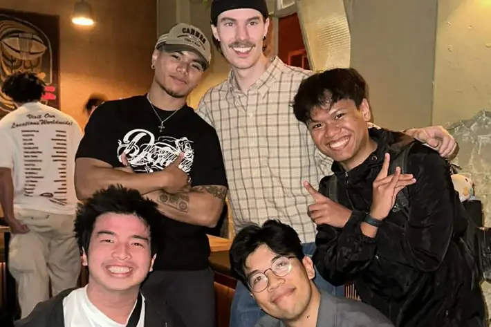 A group of five people pose together inside a casual indoor venue. Three are standing and two are crouching in front, all making playful hand signs. They are dressed in relaxed clothing, including jackets, T-shirts and caps. The background shows other patrons, warm lighting and wall décor, creating a lively social atmosphere.