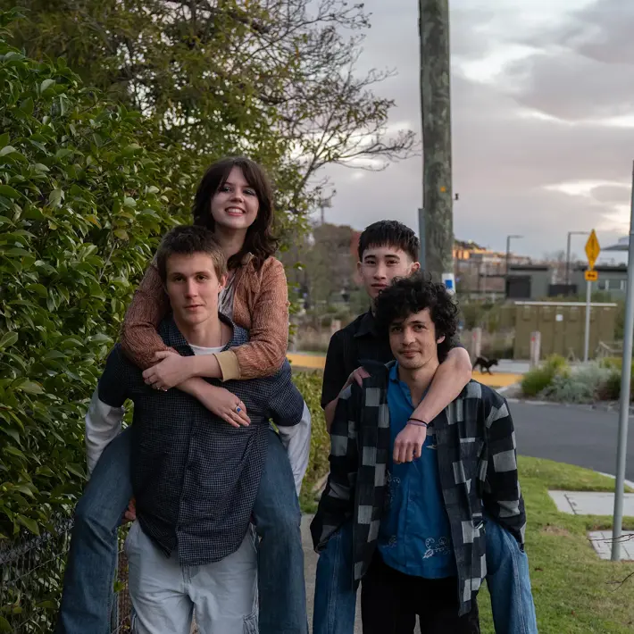 Four people are outdoors on a suburban footpath, with two giving piggyback rides to the others. The group is dressed casually in jeans, shirts, and sneakers, with one wearing a checkered overshirt and another in a patterned top. Behind them are green bushes, a street with signage, and houses in the distance under a cloudy sky. The scene feels relaxed and informal, capturing a playful moment in a neighbourhood setting.