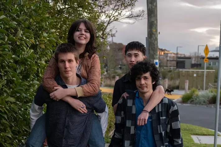 Four people are outdoors on a suburban footpath, with two giving piggyback rides to the others. The group is dressed casually in jeans, shirts, and sneakers, with one wearing a checkered overshirt and another in a patterned top. Behind them are green bushes, a street with signage, and houses in the distance under a cloudy sky. The scene feels relaxed and informal, capturing a playful moment in a neighbourhood setting.
