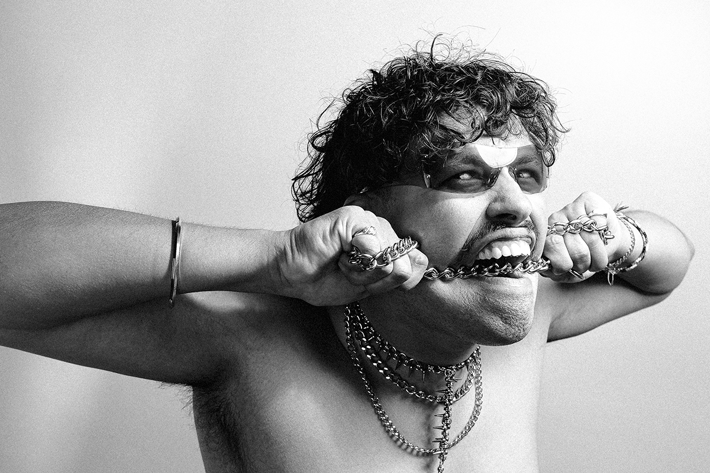 A black-and-white image of a person biting a chain, wearing layered necklaces, bracelets, and sunglasses, with an intense expression.
