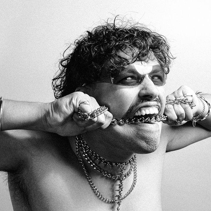 A black-and-white image of a person biting a chain, wearing layered necklaces, bracelets, and sunglasses, with an intense expression.