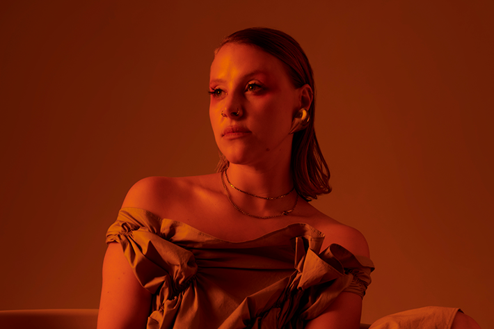 A person with shoulder-length hair sits in a moody, orange-toned setting, wearing an off-the-shoulder draped dress and plaid gloves. Their calm gaze and soft makeup, with a pop of red near the eyes, are highlighted by dramatic, warm lighting.