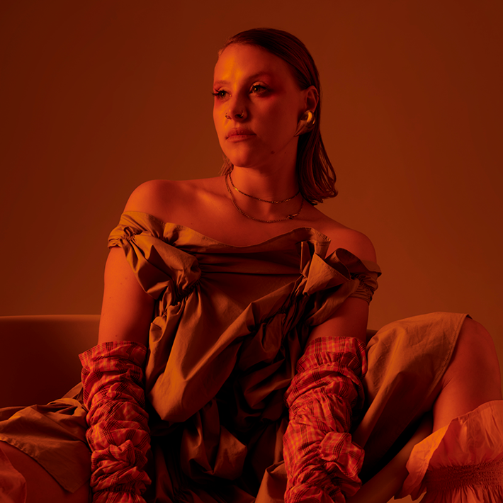 A person with shoulder-length hair sits in a moody, orange-toned setting, wearing an off-the-shoulder draped dress and plaid gloves. Their calm gaze and soft makeup, with a pop of red near the eyes, are highlighted by dramatic, warm lighting.