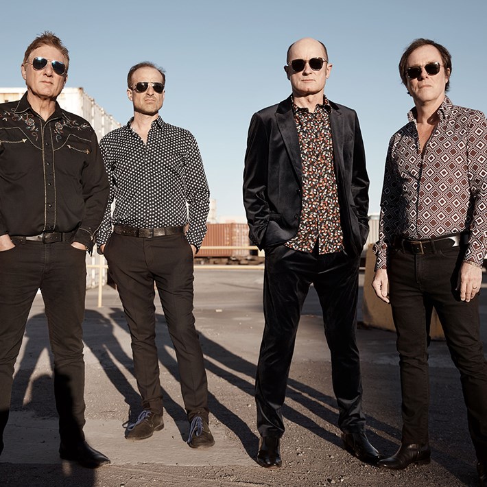 Four men standing in a shipping container yard. They are wearing dark clothing and sunglasses.