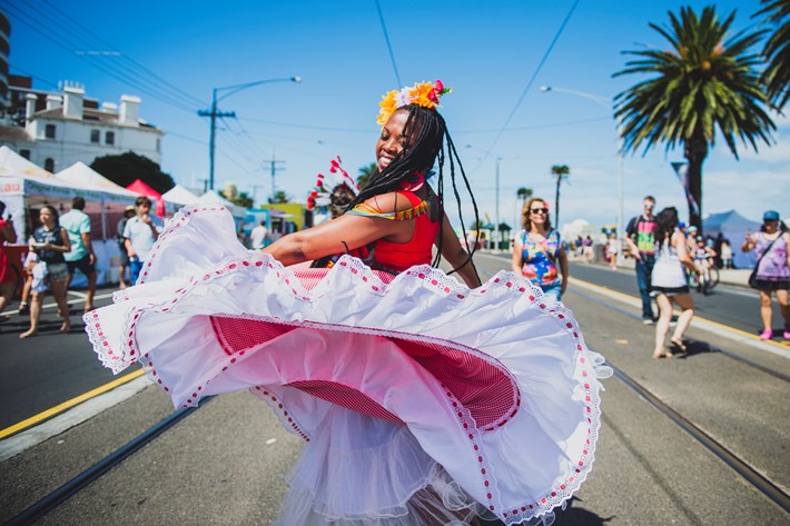 A dancer with long dark braids, a flower headpiece, a red top and a red skirt with a large white frill at the bottom is spinning around while flipping their skirt in the middle of a road surrounded by people.